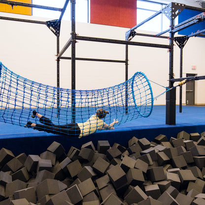 A girl climbing through the rope tunnel while suspended over a foam pit.